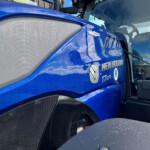 Close-up of a blue truck cab with a large metal mesh grille and raindrops on the surface