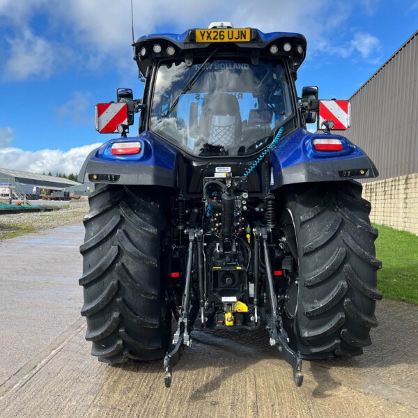 Rear view of a blue agricultural tractor with large tires and attached hitch, parked outside a farm building under a blue sky.