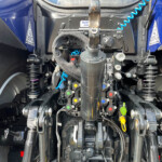 Rear tractor connection area with hydraulic hoses, cables, and metal linkages for trailer hookup; blue truck body visible on sides.