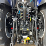 Rear view of a heavy agricultural tractor hitch with hydraulic controls, hoses, and yellow hitch component between large treads.