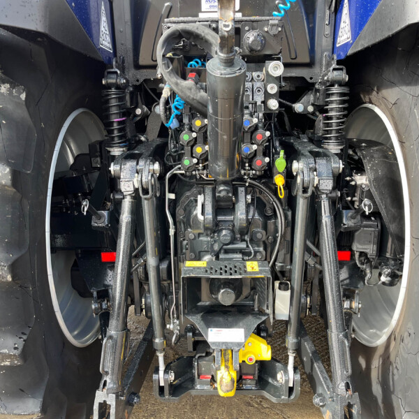 Rear view of a heavy agricultural tractor hitch with hydraulic controls, hoses, and yellow hitch component between large treads.