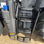 Close-up of a truck’s front steps and chassis, wet with raindrops, showing a blue reservoir cap and orange reflector at the lower left.