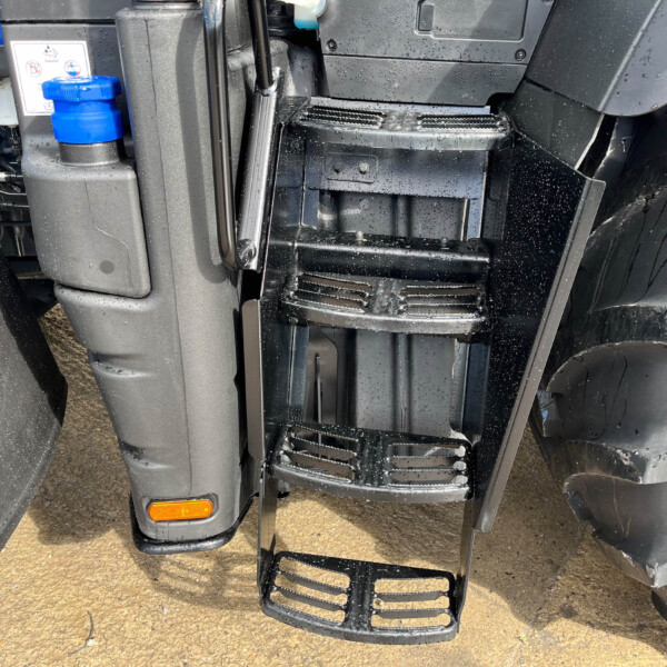 Close-up of a truck’s front steps and chassis, wet with raindrops, showing a blue reservoir cap and orange reflector at the lower left.