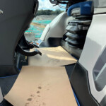 Interior of a boat cockpit with pedal controls and stacked seats, looking toward a wet deck and turquoise water outside