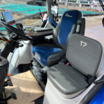 Cab interior of a construction vehicle with a blue 'Blue Power' seat, steering wheel, pedals, and control panel; construction site visible through the windshield.