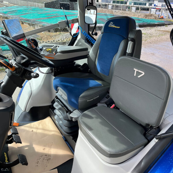 Cab interior of a construction vehicle with a blue 'Blue Power' seat, steering wheel, pedals, and control panel; construction site visible through the windshield.