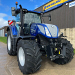Blue New Holland agricultural tractor parked outside a corrugated-metal building with a yellow 'New Holland Agriculture' sign in the background, large rear tires visible.