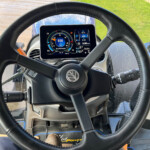 Interior view of a tractor cab with a circular digital gauge reading 850 and the steering wheel in front.