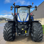 Front view of a large blue tractor with oversized tires parked in an industrial yard beside a corrugated metal building with a bright blue sky behind it.