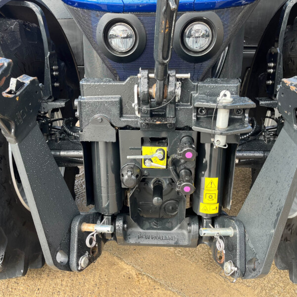 Close-up of a New Holland tractor front hitch with hydraulic mechanism, two round lights, and warning stickers on a muddy ground surface.