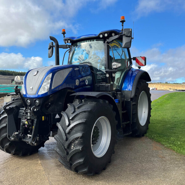 Blue New Holland tractor with oversized tires parked on a paved surface under a partly cloudy sky in a rural yard.