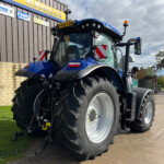 Blue agricultural tractor with large front tires parked beside a brick wall and a building with signs in the background, front cab facing the camera.