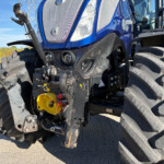 Front view of a blue tractor showing the grille, dual headlights, and large off-road tires with visible hydraulic components and metal hitch at the front.