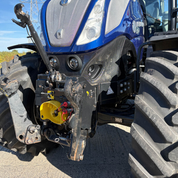 Front view of a blue tractor showing the grille, dual headlights, and large off-road tires with visible hydraulic components and metal hitch at the front.