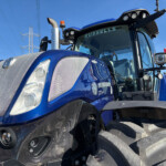 Close-up of a blue tractor cab with a chrome exhaust stack, large headlights, and the Russell logo on the windshield against a clear blue sky.