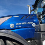 Close-up of a blue New Holland T7.300 truck cab with chrome exhaust, extra lights, and branding against a clear sky.