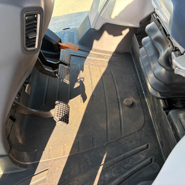 Inside a truck cab showing three pedals (clutch, brake, accelerator) and a rubber floor mat with sunlight casting shadows, near the driver's area.