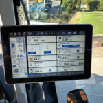 Close-up of a tablet in a tractor cab showing engine RPM, fuel, and system status panels with an outdoor yard in view behind it.