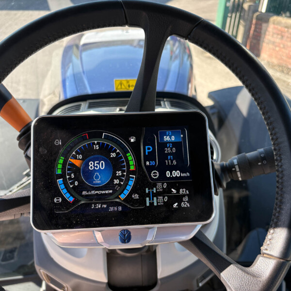 Driver's-view of a modern tractor dashboard with a circular digital gauge showing 850 (RPM) and surrounding meters, steering wheel in foreground
