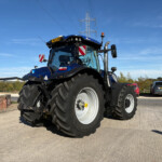 Blue New Holland tractor front view with oversized tires, hazard markers on the sides, parked in a lot under a clear blue sky.