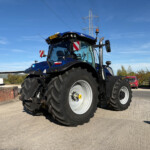 Blue New Holland tractor seen from the front-left in a parking lot, with a clear blue sky and power-line tower in the background.
