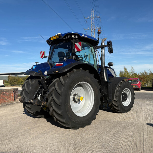 Blue New Holland tractor seen from the front-left in a parking lot, with a clear blue sky and power-line tower in the background.
