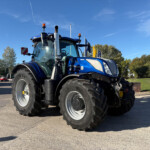 Close-up of a blue New Holland tractor parked outdoors, showing its cab, oversized tires, and exhaust against a clear blue sky.