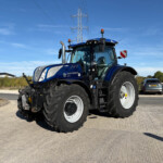 Blue New Holland tractor from a low angle, with tall power-line tower in the background against a clear blue sky.