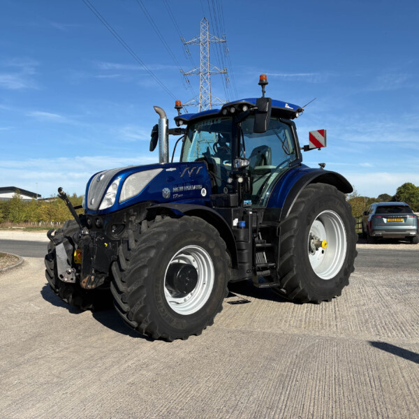 Blue New Holland tractor from a low angle, with tall power-line tower in the background against a clear blue sky.