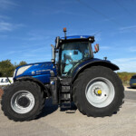 Blue New Holland tractor (T7.300) close-up with large front wheels on a paved lot under a clear blue sky, power lines in the background.