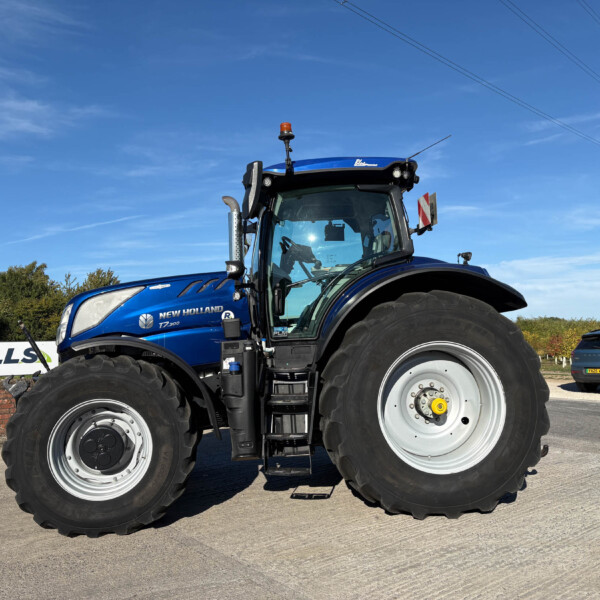 Blue New Holland tractor (T7.300) close-up with large front wheels on a paved lot under a clear blue sky, power lines in the background.