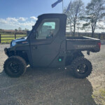 Rugged Polaris Ranger side-by-side ATV on a gravel lot with a wooden fence and rural fields in the background.