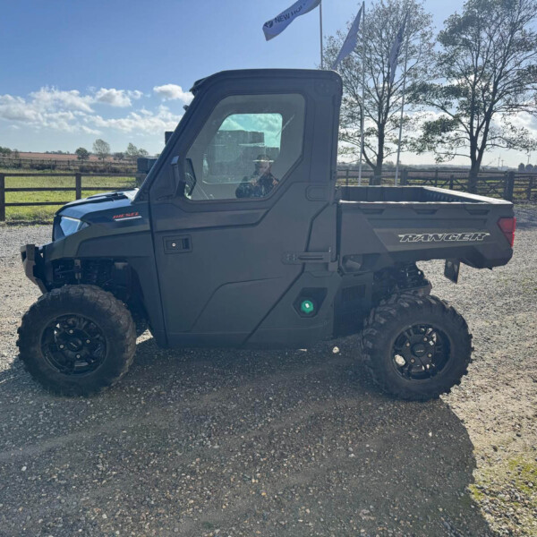 Rugged Polaris Ranger side-by-side ATV on a gravel lot with a wooden fence and rural fields in the background.