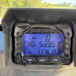 Close-up of a Polaris digital gauge cluster; shows 0 mph, fuel icon, and control buttons, with an outdoor landscape in the background.