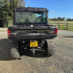 Rear view of a black Polaris off-road vehicle with large tires parked on a gravel driveway; a rural field and wooden fence are in the background.