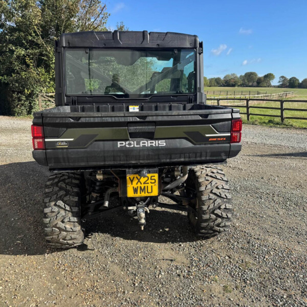 Rear view of a black Polaris off-road vehicle with large tires parked on a gravel driveway; a rural field and wooden fence are in the background.
