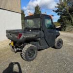 Black Polaris Ranger side cab parked on a gravel driveway beside a light-colored wall and wooden gate, with trees in the background.