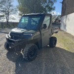 Gray side-by-side utility vehicle parked on a gravel lot with trees in the background and a white wall/building to the right.