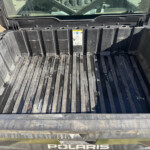 Empty black plastic bed liner in a Polaris off-road vehicle, with a dirt-streaked floor and a small warning label on the side wall