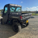Black Polaris Ranger utility vehicle parked on a gravel lot, with a rural fence and hay bale stacks in the background under a blue sky.