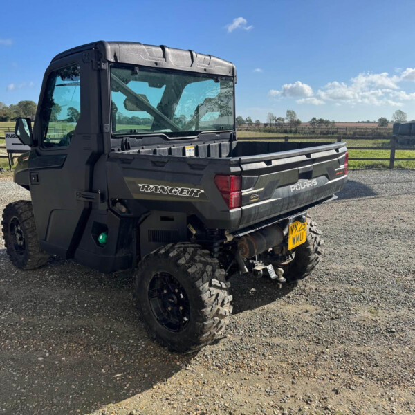 Black Polaris Ranger utility vehicle parked on a gravel lot, with a rural fence and hay bale stacks in the background under a blue sky.
