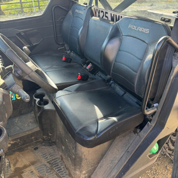 Interior of a Polaris Ranger UTV with two front seats, seat belts, and a muddy floor; outdoor rural scene visible through the window.