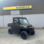 All-terrain off-road vehicle with large tires parked on gravel in front of a gray corrugated wall with a yellow/blue Russell's sign above.