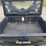 Rear view of a black Polaris utility vehicle with its cargo bed open, showing the cab and windshield inside on a gravel driveway.