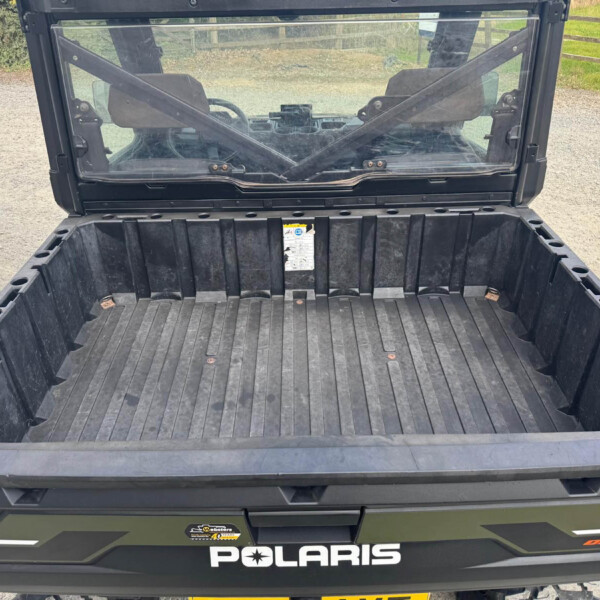 Rear view of a black Polaris utility vehicle with its cargo bed open, showing the cab and windshield inside on a gravel driveway.