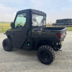 Black off-road utility vehicle parked on a gravel lot with a field in the background and stacked wrapped hay bales to the right side of the image.