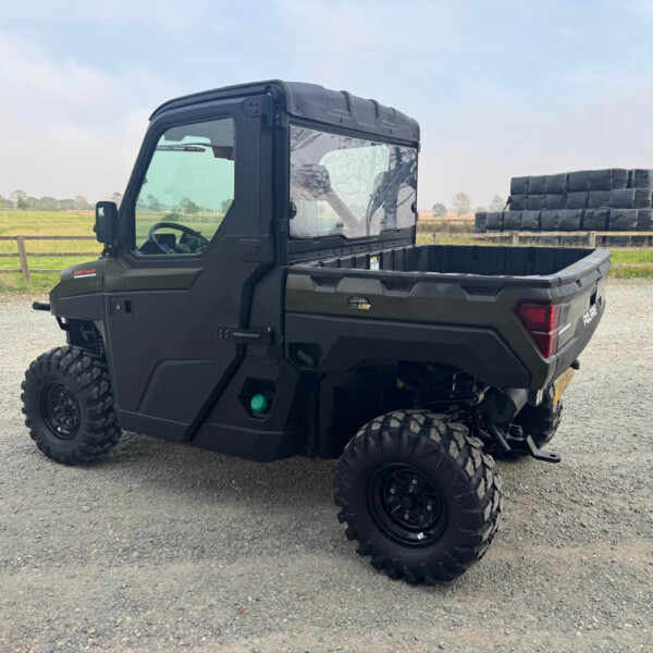 Black off-road utility vehicle parked on a gravel lot with a field in the background and stacked wrapped hay bales to the right side of the image.