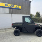 Black off-road utility vehicle with large tires parked in a gravel lot beside a grey industrial building with a Russell's sign.