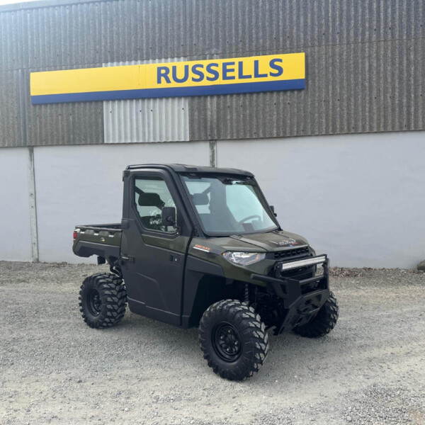 All-terrain off-road vehicle with large tires parked on gravel in front of a gray corrugated wall with a yellow/blue Russell's sign above.