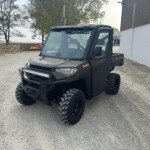 Front three-quarter view of a dark green off-road utility vehicle with large knobby tires and a roof, parked on a gravel lot near a barn.